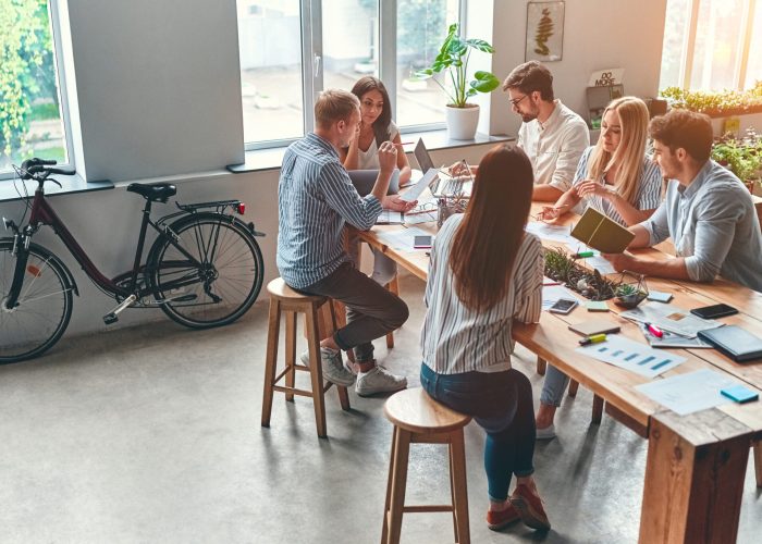 Eine Gruppe Menschen sitzt an einem Holztisch in einem offenen Co-Working Space und unterhalten sich über Arbeitsergebnisse